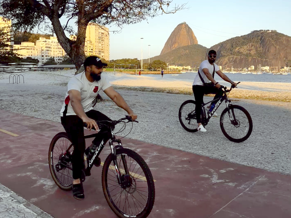 Two men ride black mountain bikes along a cycleway with Rio's Sugarloaf mountain the background.